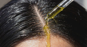 macro close-up of a glass pipette dripping yellowish earthy green olive oil onto a wet black-haired scalp in direct sunlight

